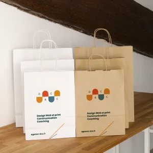 Sacs promotionnels personnalisés en papier blancs et bruns imprimés avec le logo et les services de l’agence, placés sur une étagère en bois sous une poutre rustique.”
